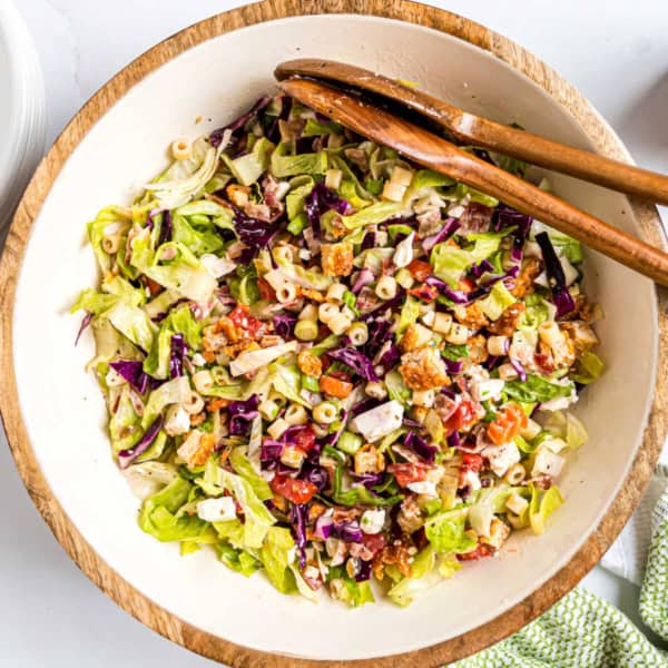 A bowl of chopped salad with pasta, lettuce, tomatoes, croutons, and purple cabbage, with wooden tongs.