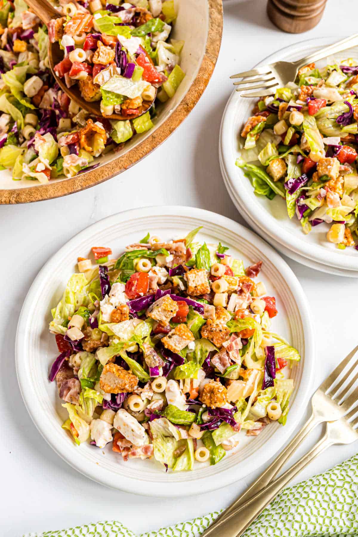 Two plates and a bowl of colorful chopped salad with veggies, pasta, and croutons, served with forks.