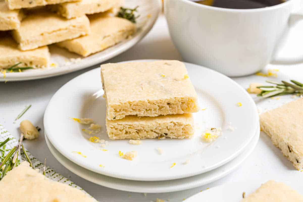Two square shortbread cookies stacked on a white plate, with a cup and more cookies in the background.