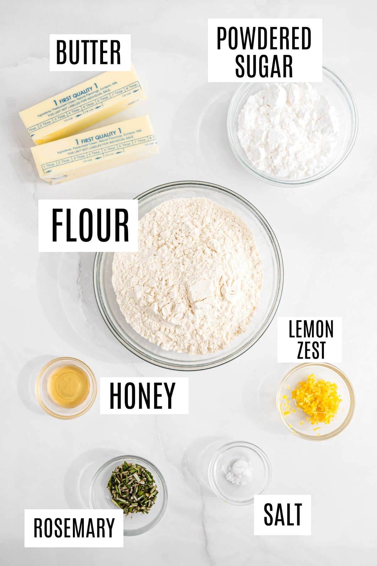 Baking ingredients on a counter: butter, powdered sugar, flour, lemon zest, honey, rosemary, and salt in bowls.