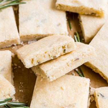 Close-up of square slices of crumbly shortbread with rosemary sprigs on a brown surface.