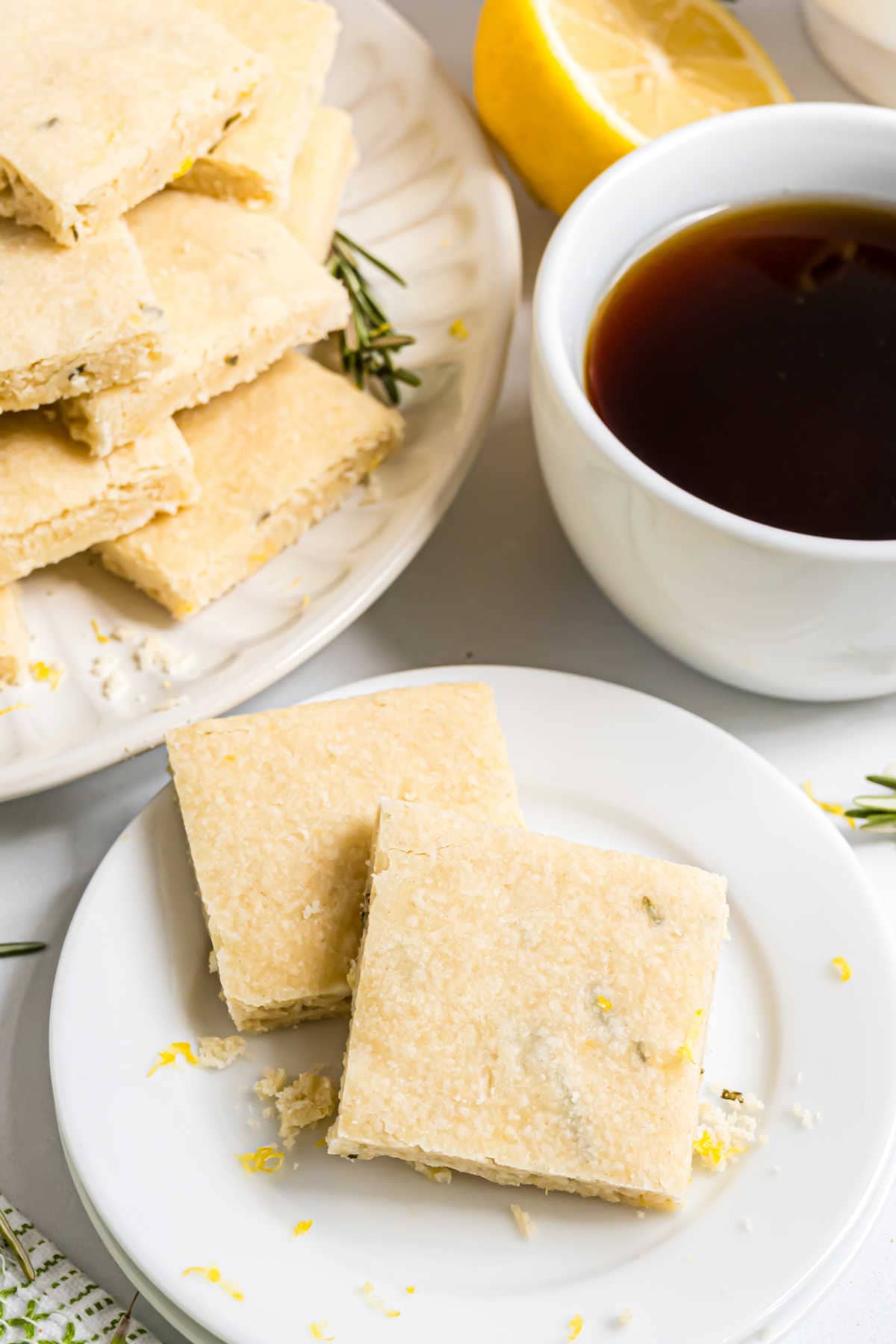 Two lemon shortbread squares on a plate, with a cup of coffee and more shortbread in the background.