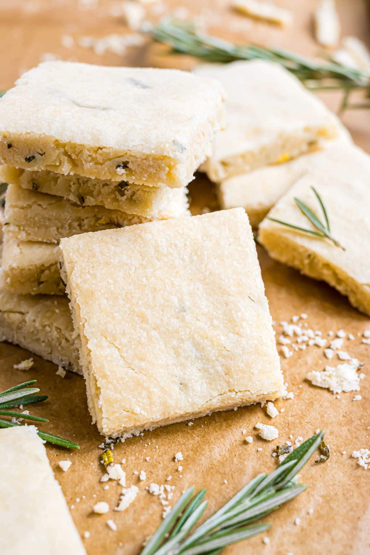 Square rosemary shortbread cookies stacked and scattered on parchment paper with rosemary sprigs nearby.