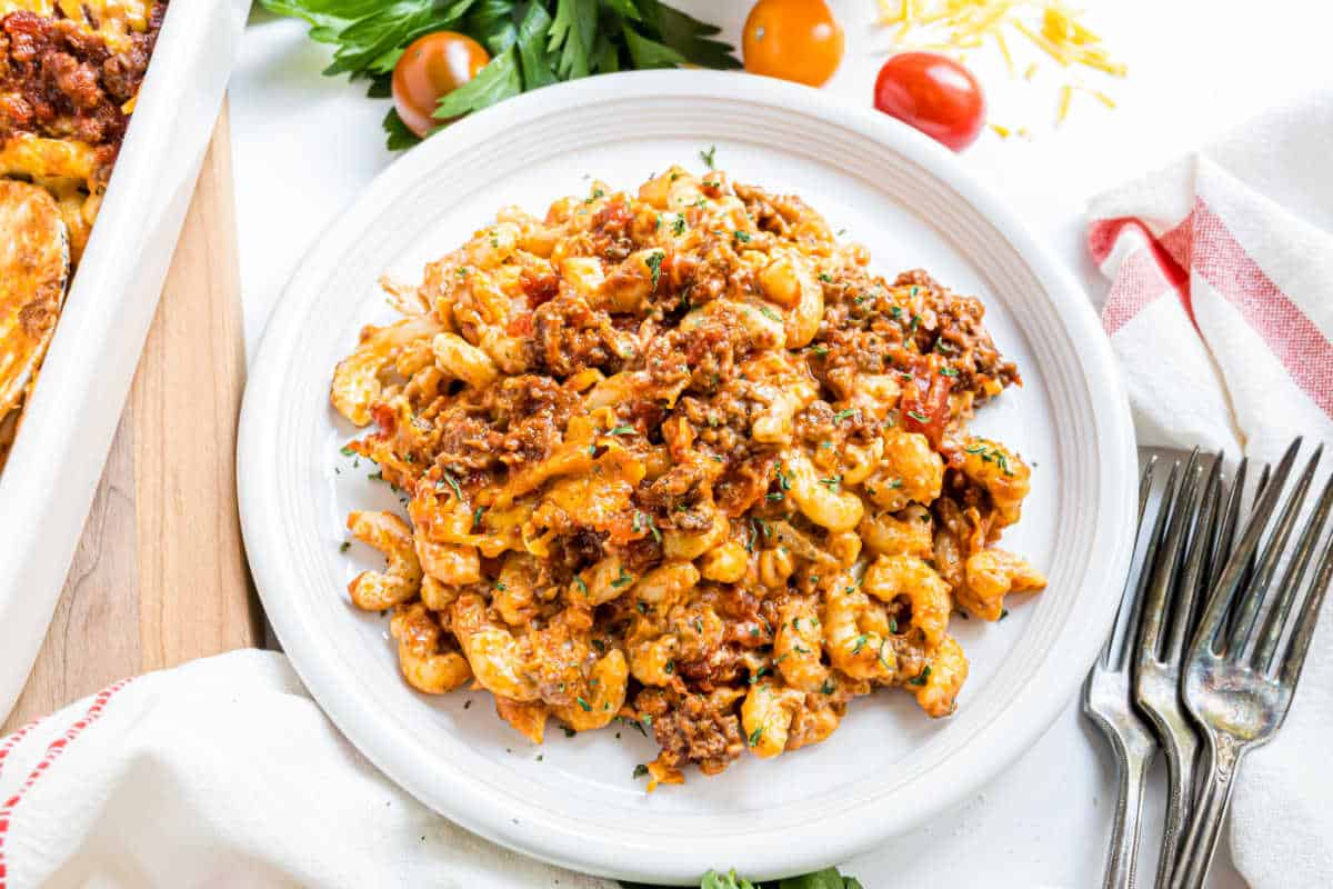 A white plate of cheesy baked pasta with ground meat and tomato sauce, garnished with herbs.