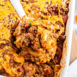 Cheesy ground beef pasta casserole being served from a white baking dish with a spoon.