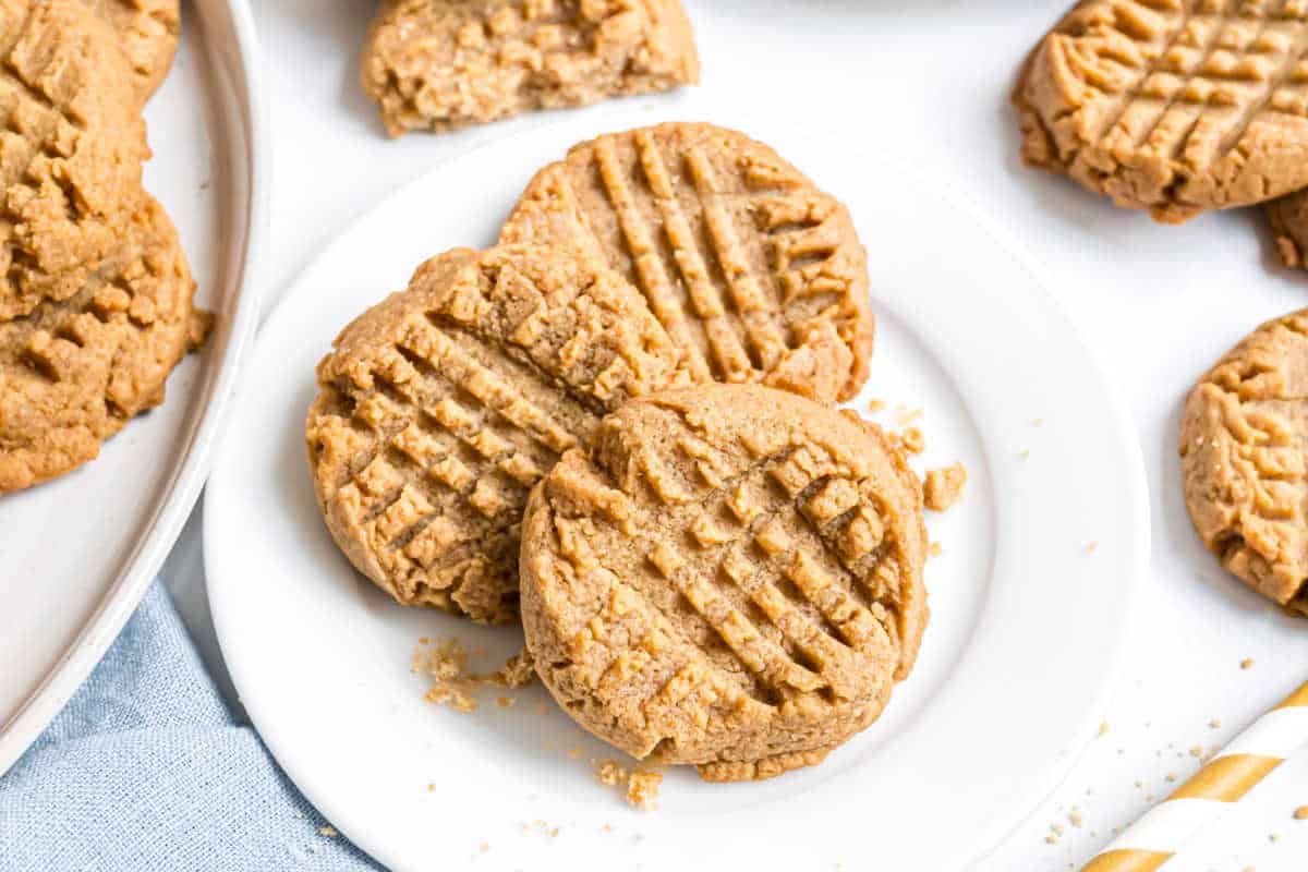 Three peanut butter cookies with fork marks on a white plate, surrounded by more cookies and crumbs.