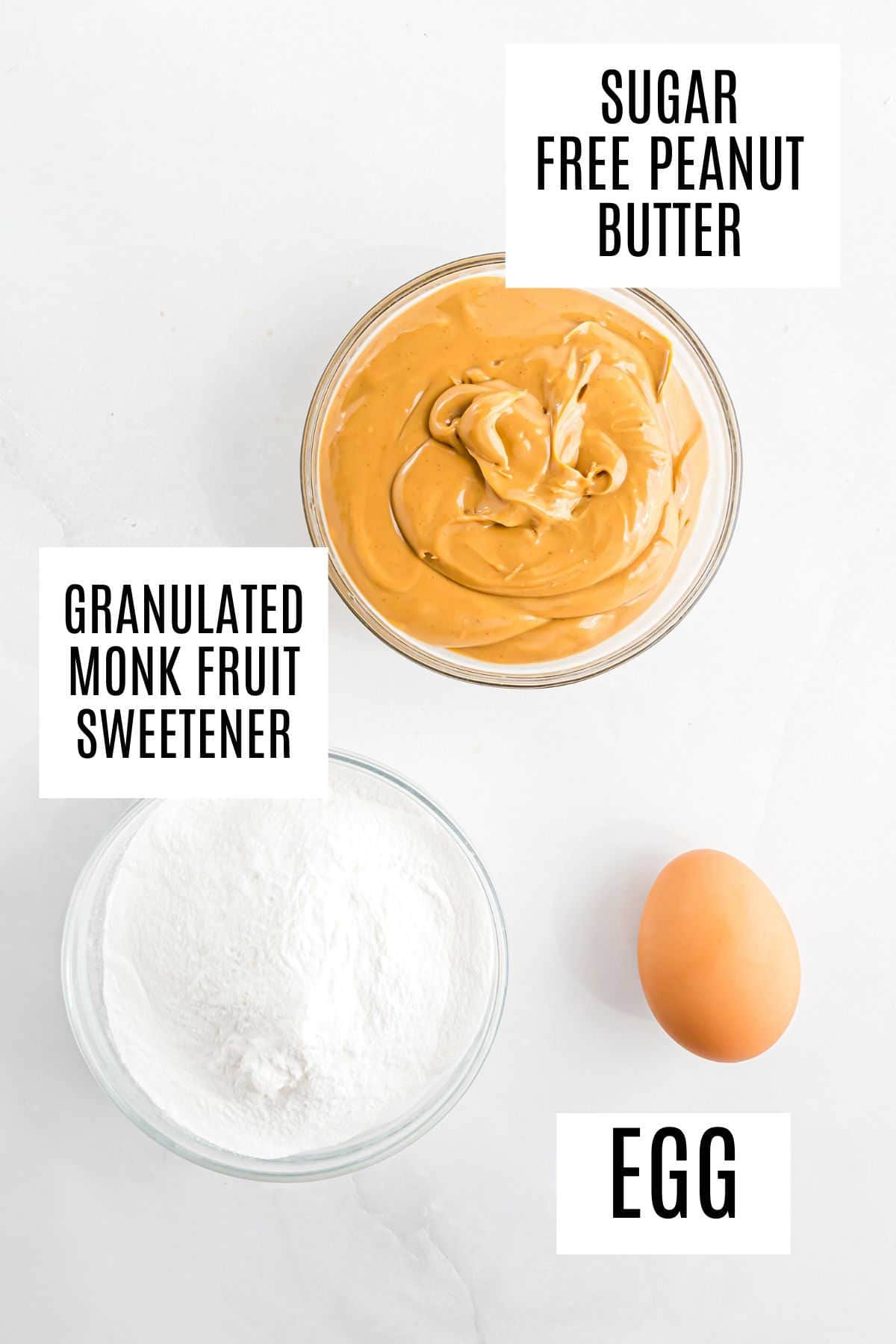 A bowl of sugar-free peanut butter, a bowl of granulated monk fruit sweetener, and a whole egg on a white surface.