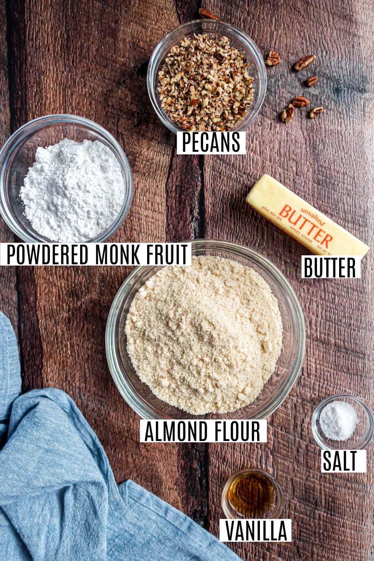 Labeled ingredients on a wooden table: pecans, powdered monk fruit, butter, almond flour, salt, and vanilla.