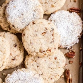 A close-up of pecan cookies, some dusted with powdered sugar, on a baking tray with scattered pecan pieces.