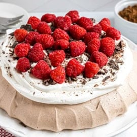 Chocolate pavlova topped with whipped cream, fresh raspberries, and chocolate shavings on a white plate.