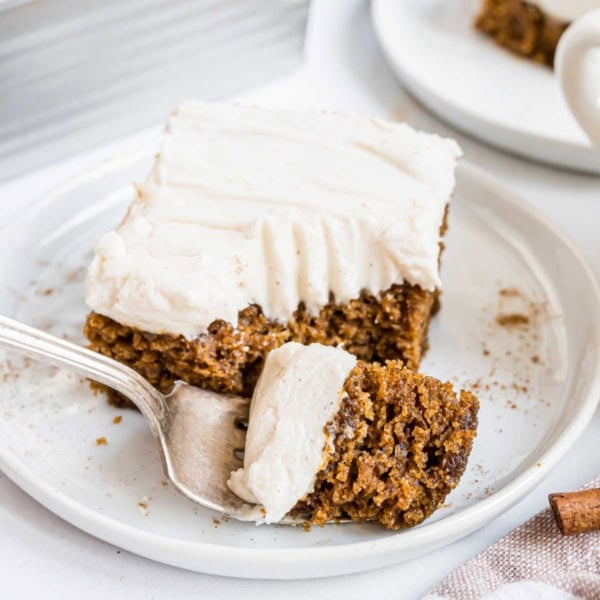 A fork cutting into a slice of spiced cake with white frosting on a white plate.