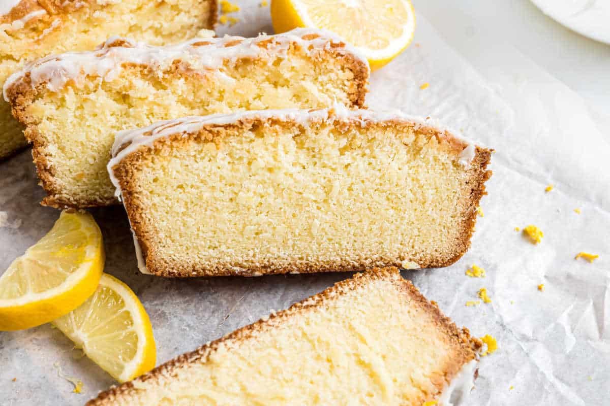 Slices of glazed lemon pound cake with lemon wedges on parchment paper.
