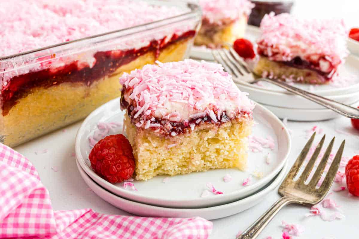 A slice of cake with raspberry jam and pink coconut flakes on a plate, beside fresh raspberries and a fork.