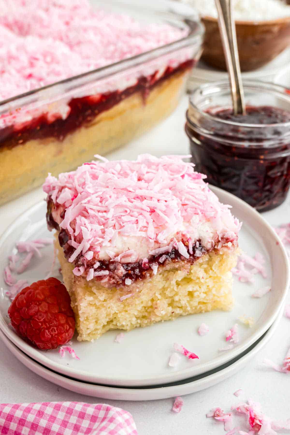 A slice of cake topped with pink coconut flakes and jam sits on a plate with a raspberry beside it.
