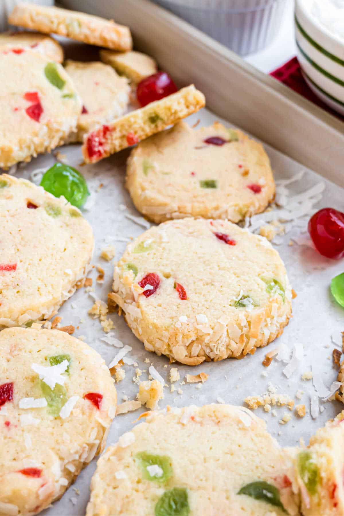 Round, sliced Santa's Whiskers cookies with red and green candied fruit and coconut rest on a baking sheet, some cookies charmingly broken.