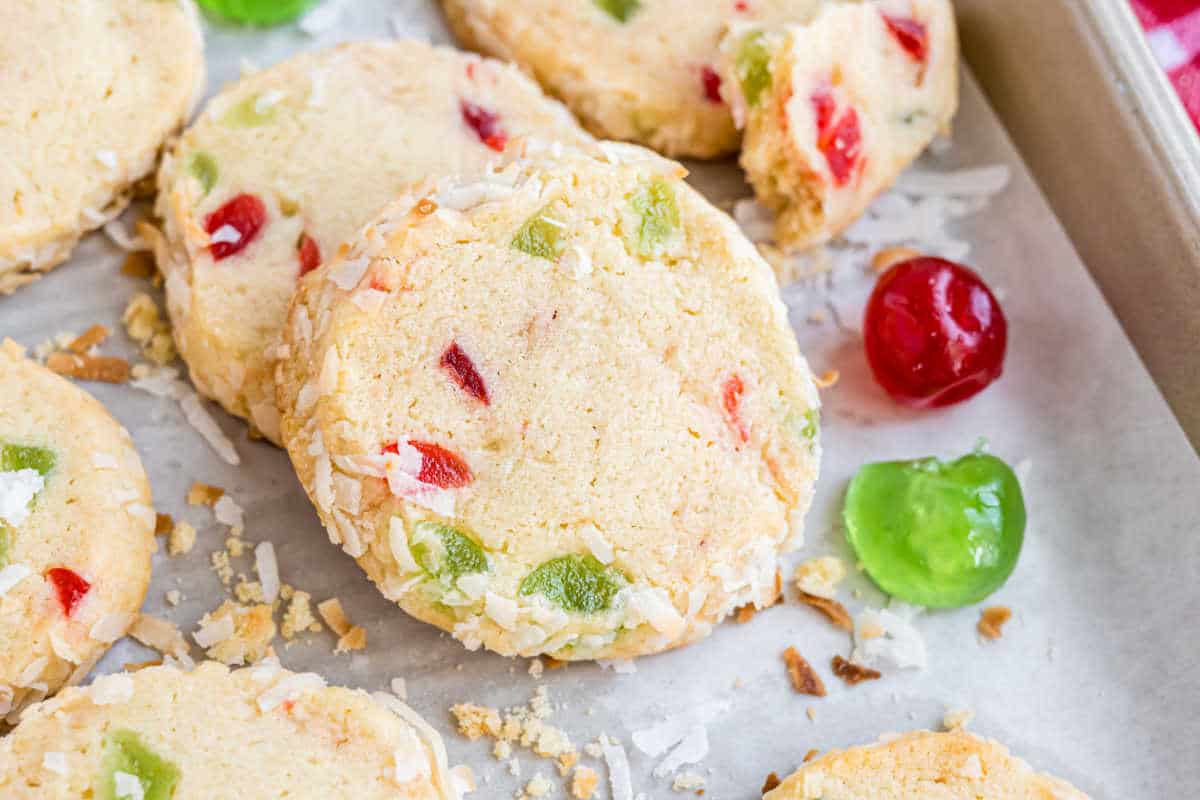 Round shortbread santa's whiskers cookies with red and green candied fruit pieces and shredded coconut on a baking sheet.