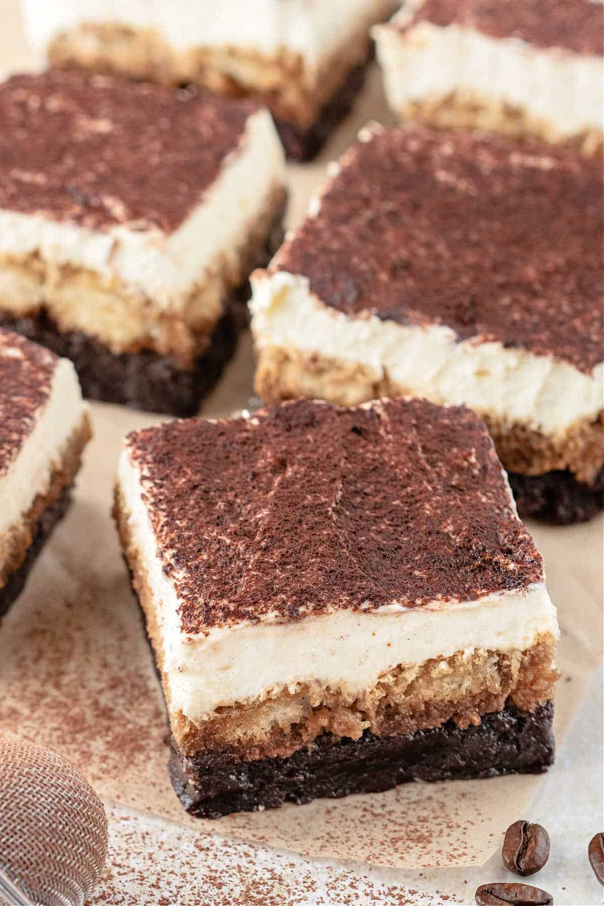 Close-up of tiramisu dessert bars with creamy layers and cocoa powder dusted on top, arranged on parchment paper.