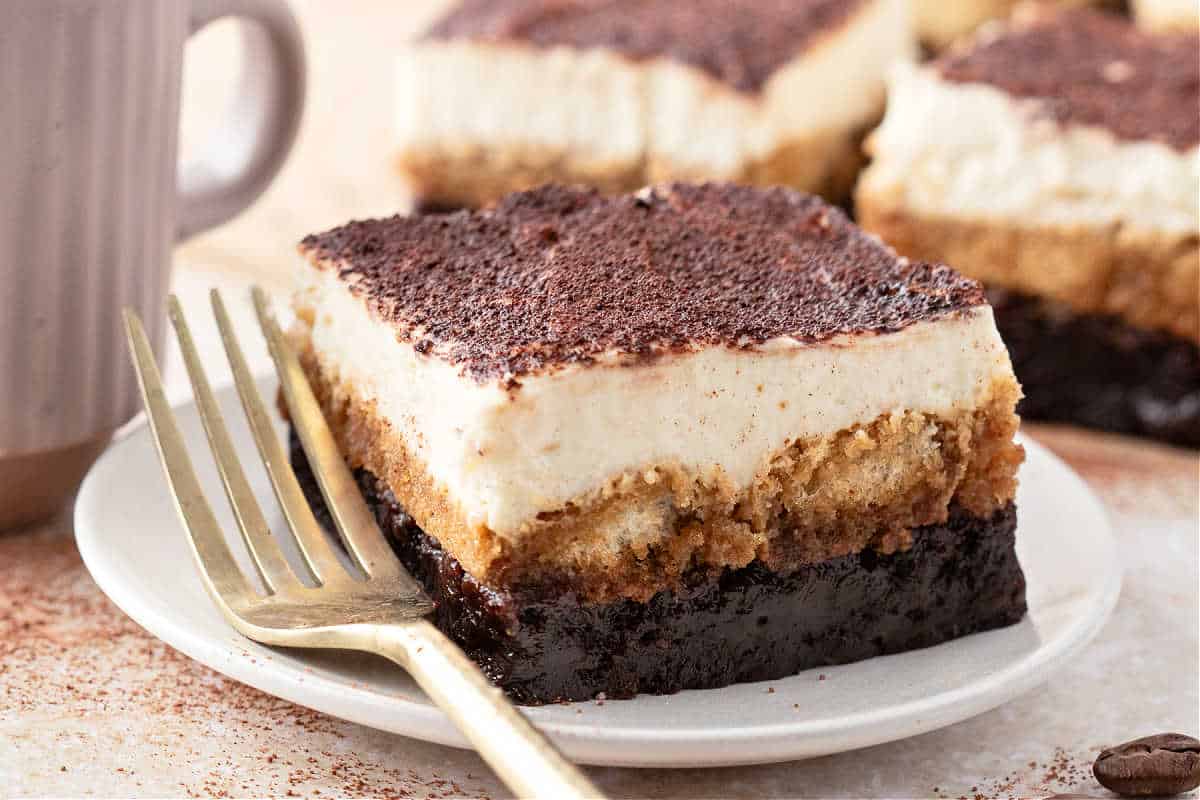 A slice of tiramisu on a plate with a gold fork, topped with cocoa powder, next to a mug.