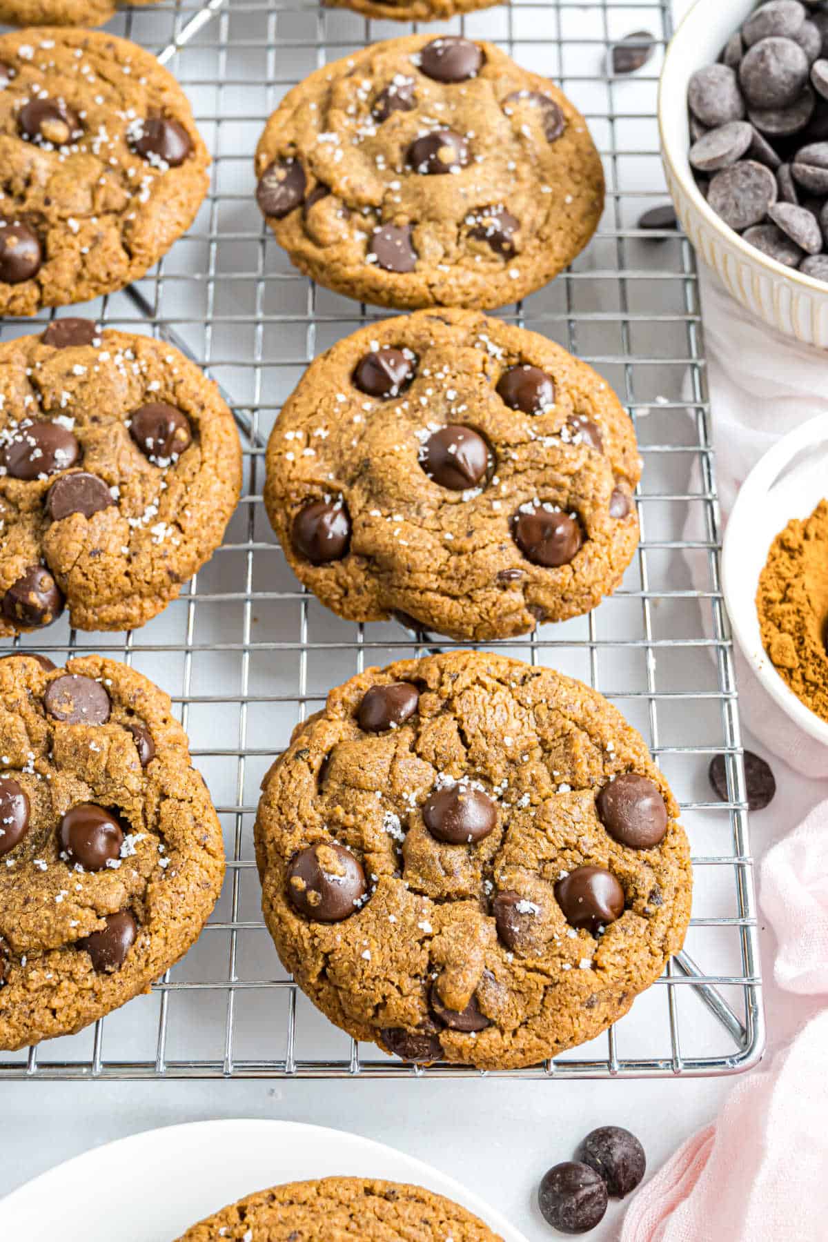Chocolate chip cookies on a cooling rack, with bowls of chocolate chips and cocoa powder nearby.