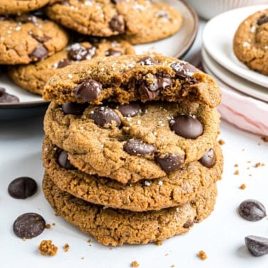 Three chocolate chip cookies stacked, topped with sea salt, with one broken in half on top.