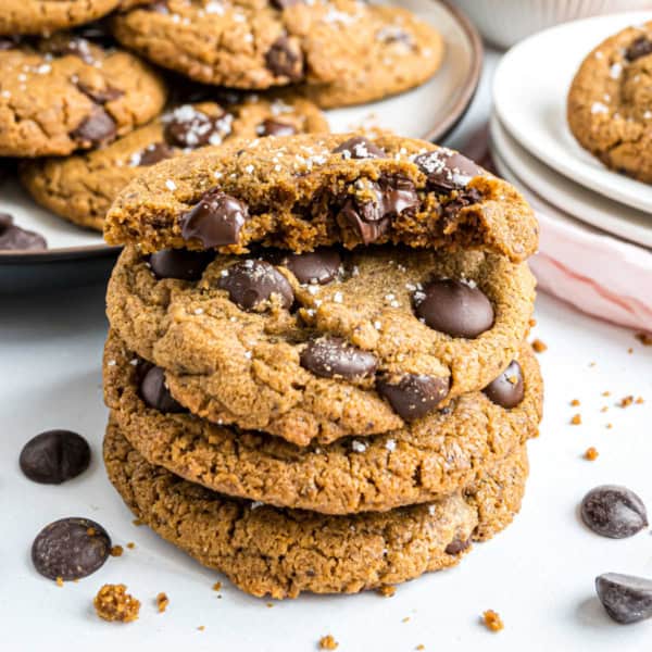 Three chocolate chip cookies stacked, topped with sea salt, with one broken in half on top.