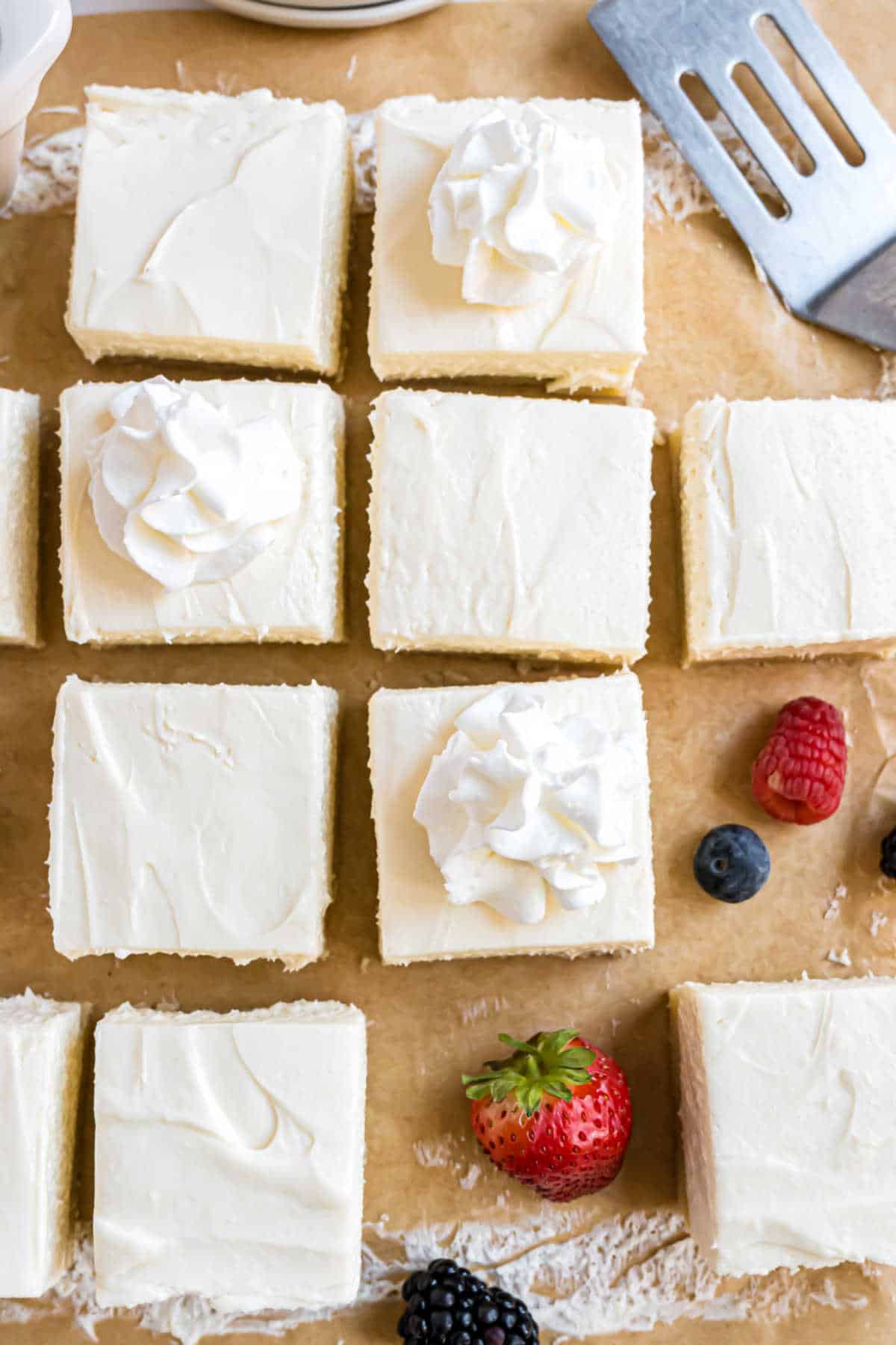 Squares of cheesecake with whipped cream on top, surrounded by fresh berries and a metal spatula.