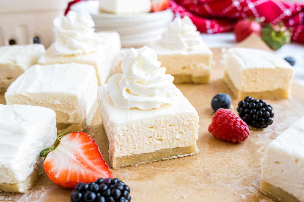 Creamy dessert bars topped with whipped cream, surrounded by fresh berries on parchment paper.
