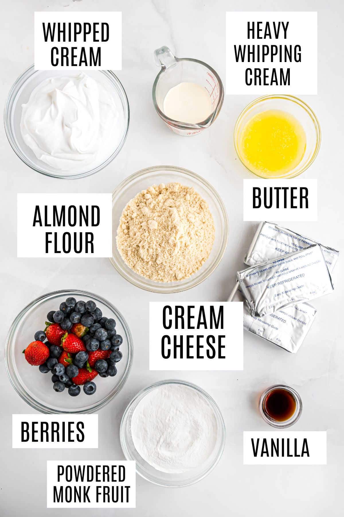 Top view of labeled ingredients in bowls: whipped cream, heavy whipping cream, butter, almond flour, cream cheese, berries, powdered monk fruit, vanilla.