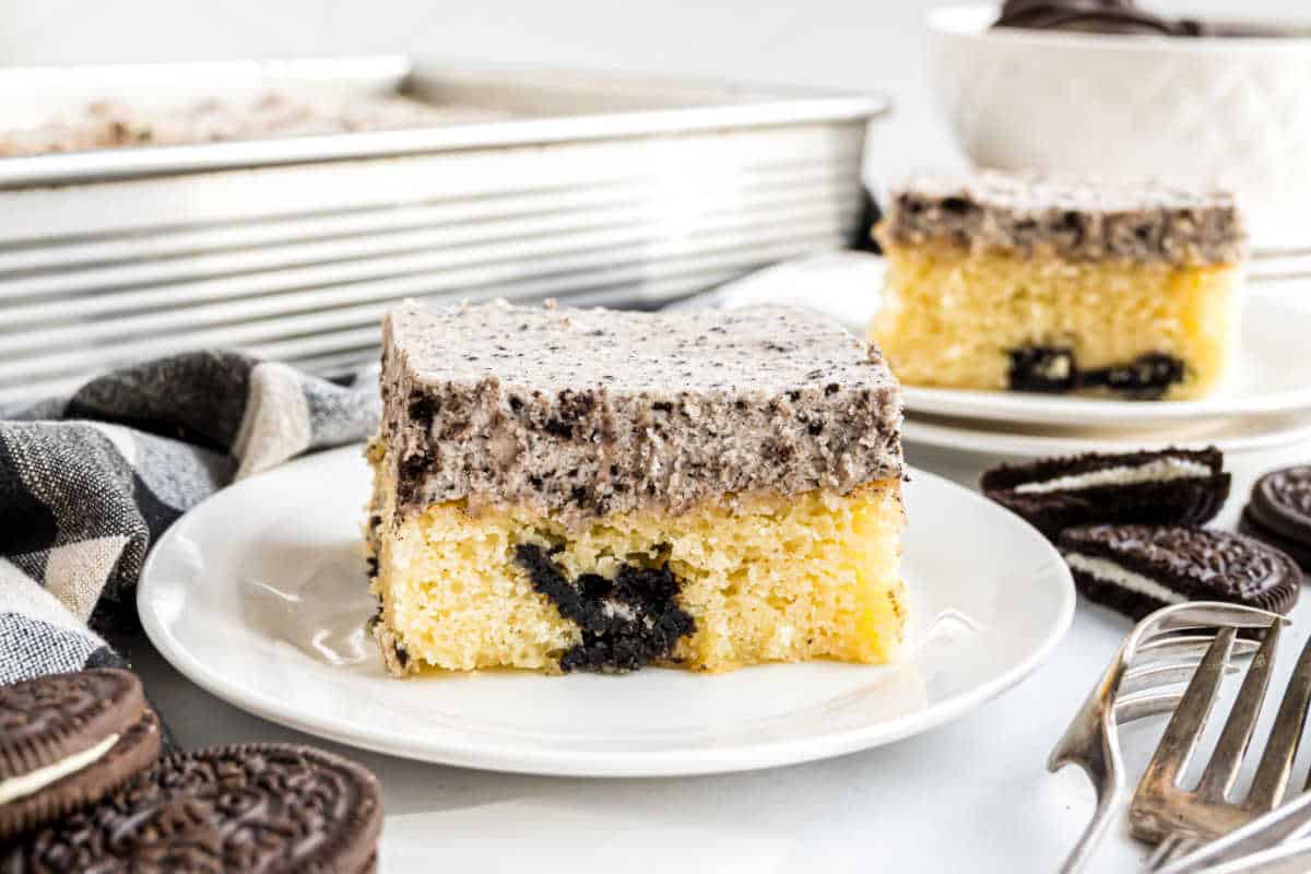 A slice of yellow cake with cookies and cream frosting on a white plate, with cookies nearby.