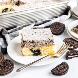 A slice of cookies and cream cake on a plate with a fork, surrounded by chocolate sandwich cookies.