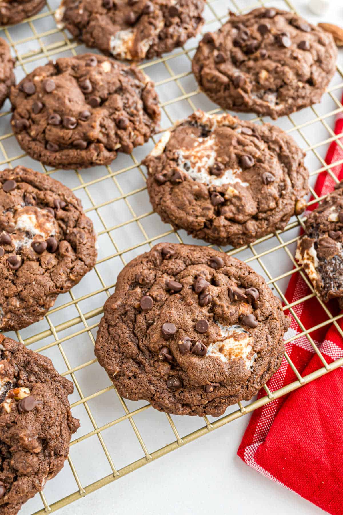 Chocolate cookies with chocolate chips and marshmallow bits on a cooling rack next to a red kitchen towel.