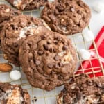 Chocolate cookies with marshmallows and chocolate chips cooling on a wire rack beside a red checkered napkin.