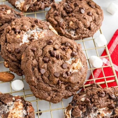 Chocolate cookies with marshmallows and chocolate chips cooling on a wire rack beside a red checkered napkin.