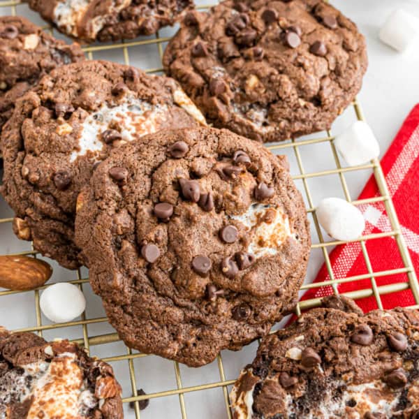 Chocolate cookies with marshmallows and chocolate chips cooling on a wire rack beside a red checkered napkin.