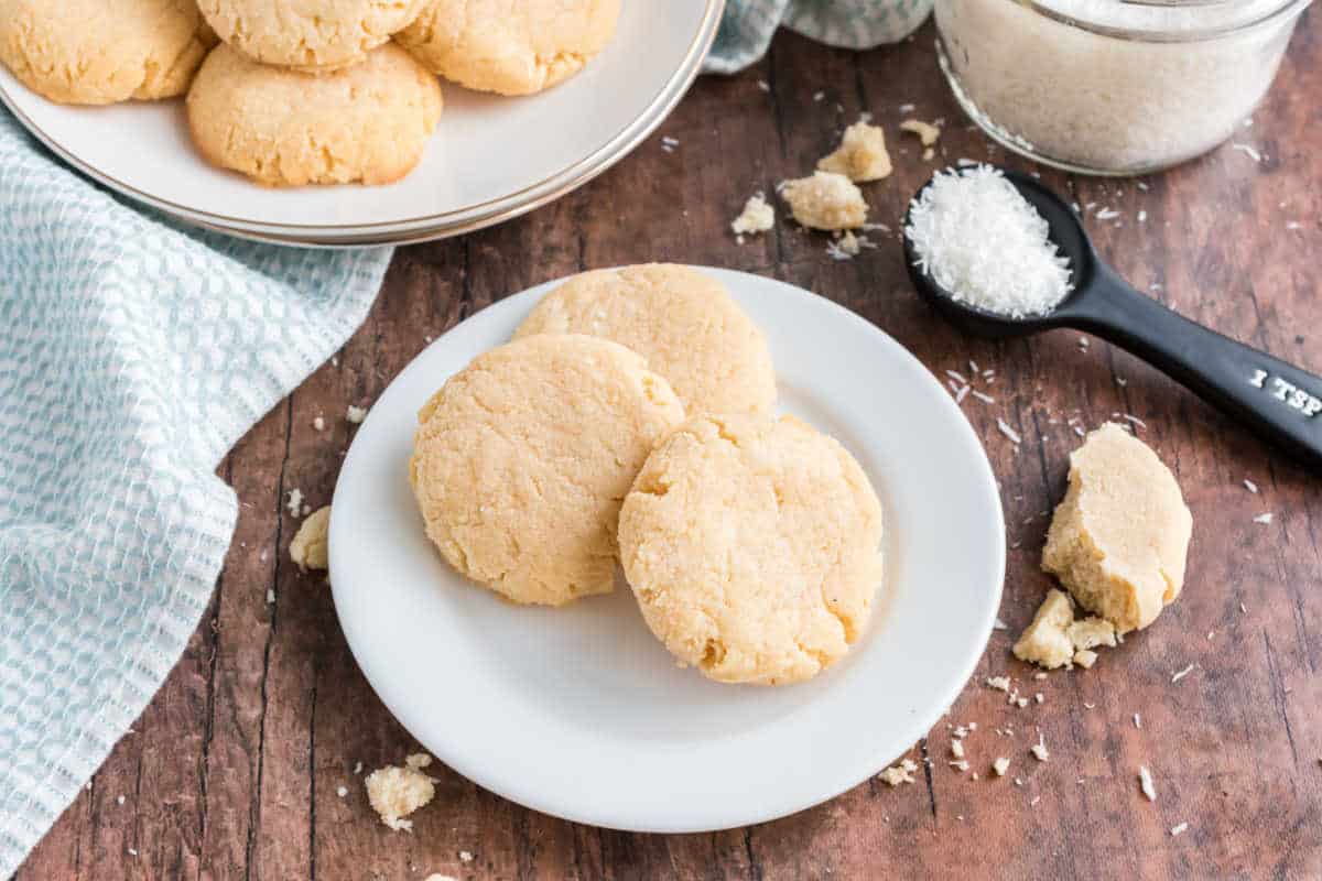 Three coconut cookies on a white plate with coconut flakes and cookie crumbs on a wooden table.