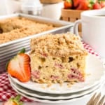 A slice of strawberry crumb cake on a plate with a fresh strawberry and a cup of coffee beside it.
