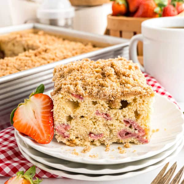 A slice of strawberry crumb cake on a plate with a fresh strawberry and a cup of coffee beside it.