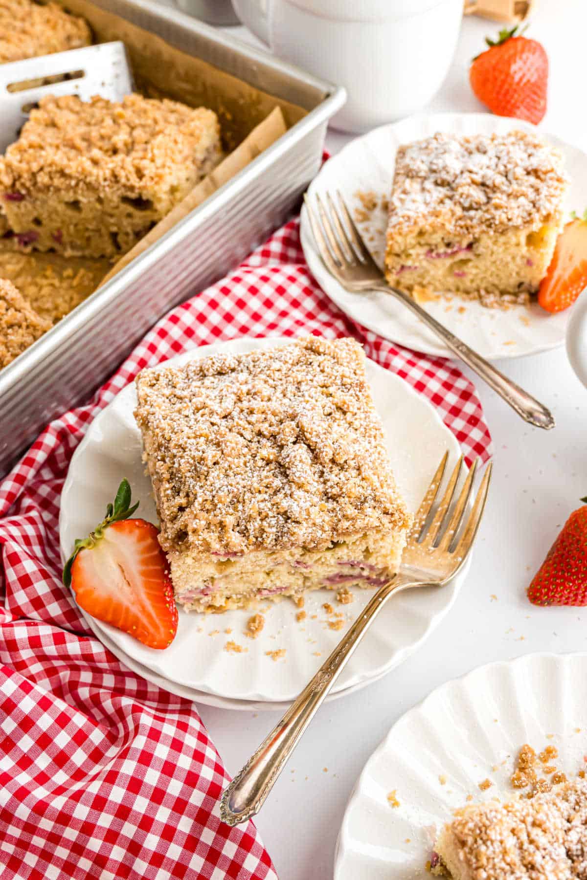 A slice of crumb-topped coffee cake with strawberries on a white plate, fork, and red-checkered napkin.
