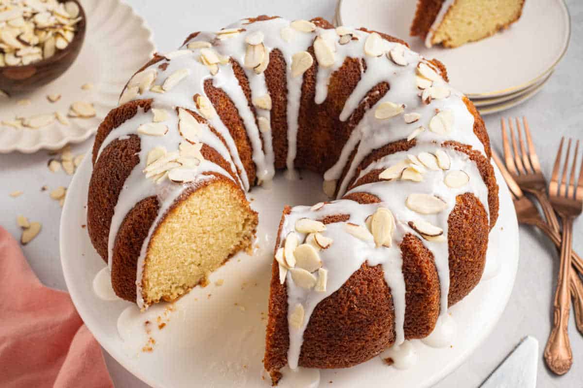 A glazed Bundt cake with sliced almonds on top, missing one slice, sits on a white cake stand.