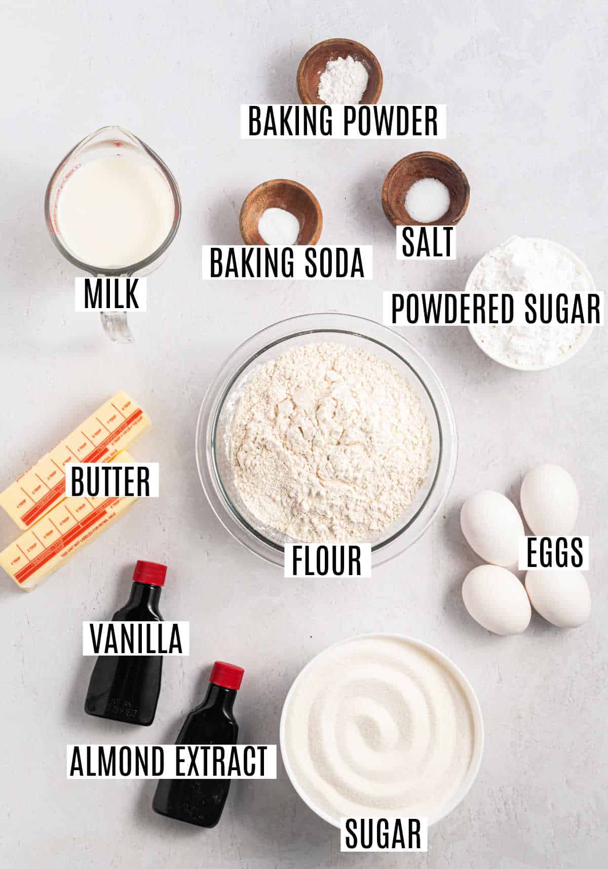 Ingredients for baking including flour, sugar, eggs, butter, milk, vanilla, almond extract, and leaveners on a table.