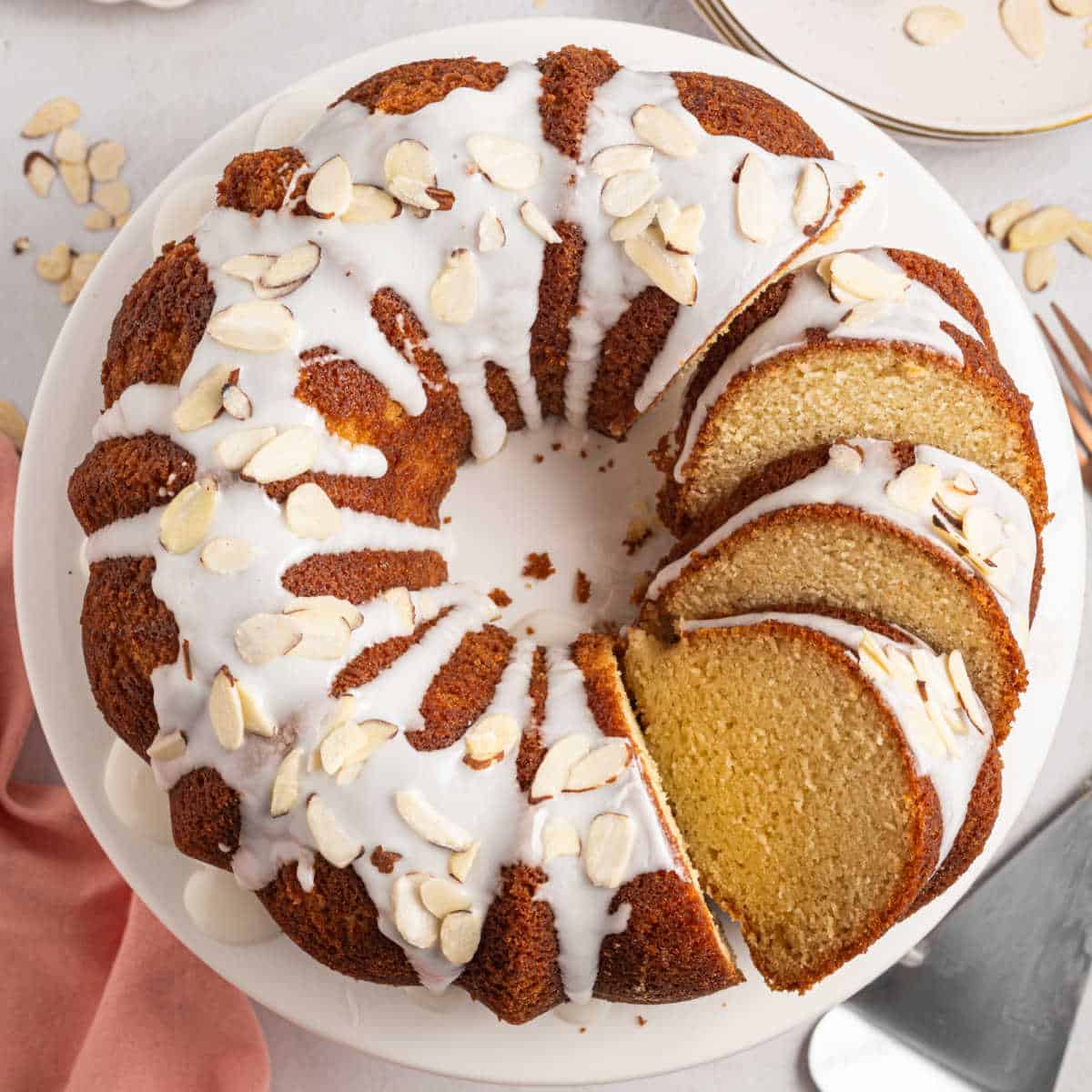Bundt cake with white icing and almond slices, partially sliced, on a white plate.