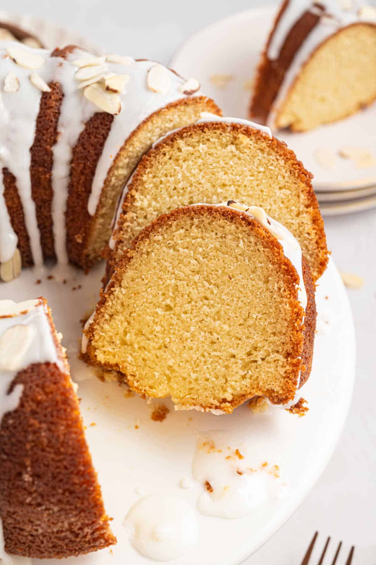 A close-up of sliced bundt cake with white glaze and almond slivers on a white plate.