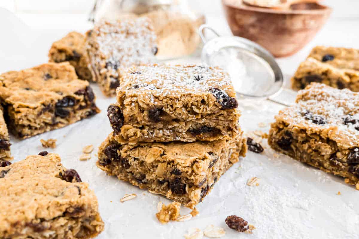 Oatmeal raisin bars stacked on parchment, dusted with powdered sugar, with oats and a sifter in the background.