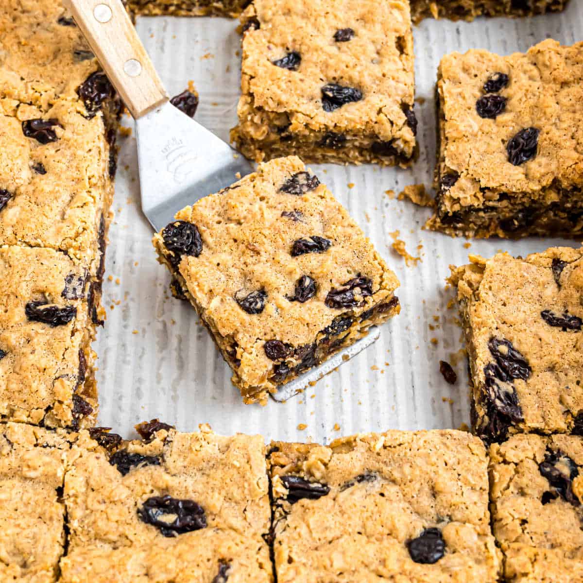 Oatmeal raisin bars cut into squares, with one piece being lifted by a spatula from a baking tray.