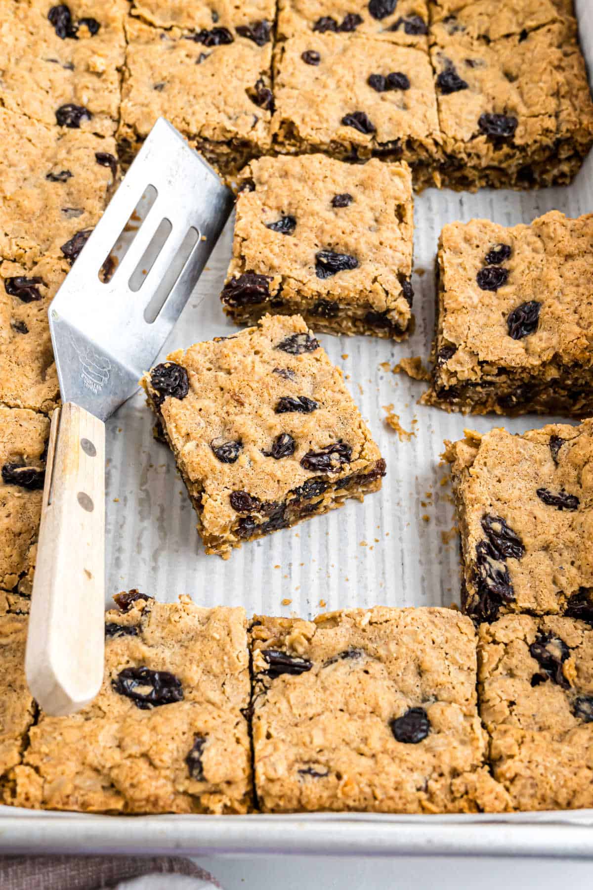 A baking pan of oat bars with raisins, some cut into squares, and a spatula resting on the bars.