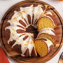 A glazed bundt cake with orange zest, sliced on a wooden platter.