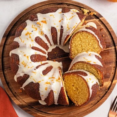 A glazed bundt cake with orange zest, sliced on a wooden platter.