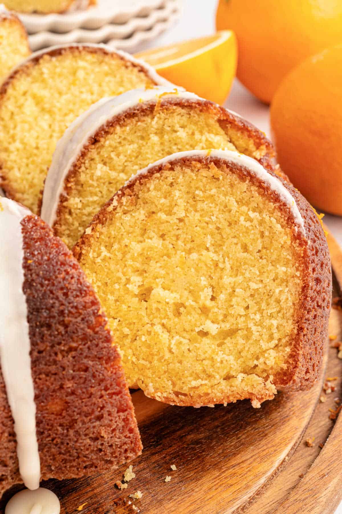 Close-up of sliced orange bundt cake with white glaze on a wooden board, fresh oranges in the background.