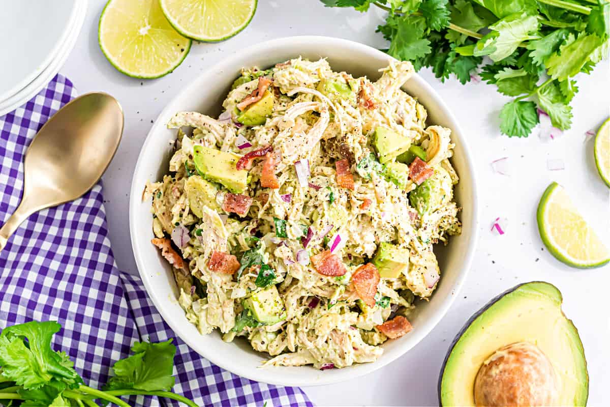 Bowl of chicken salad with avocado, bacon, and herbs, surrounded by lime, cilantro, and a gold spoon.