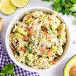 A bowl of chicken salad with avocado, red onion, bacon, and herbs, surrounded by lime slices and cilantro.